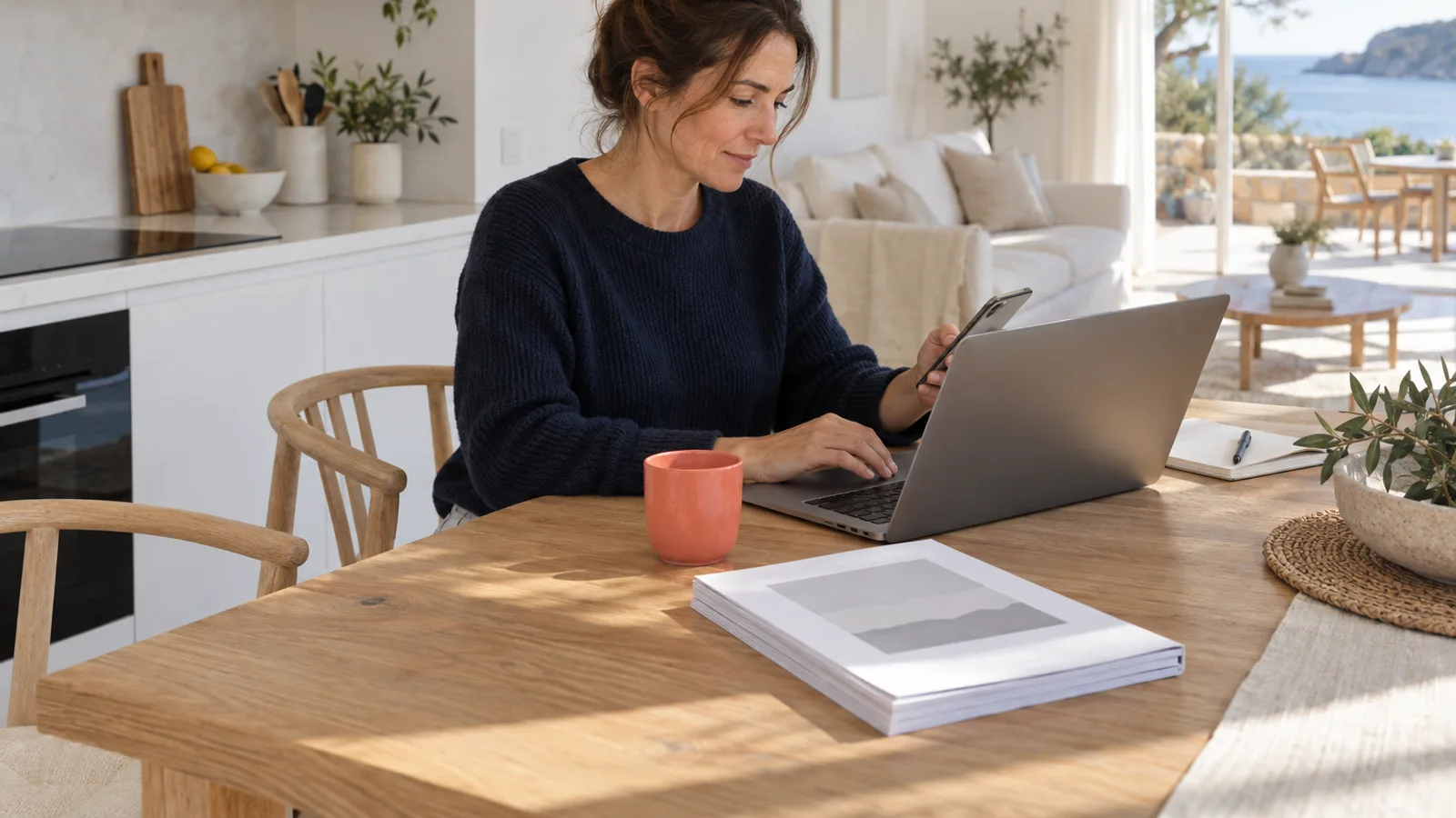 Host at a kitchen table handling guest messages on a laptop and phone next to a printed house manual